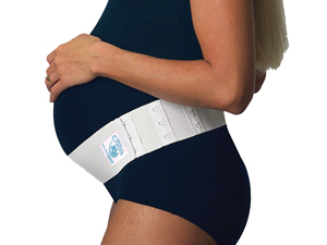 A very pregnant woman wearing black is standing up and holding the top of her stomach while wearing a white prenatal cradle strap