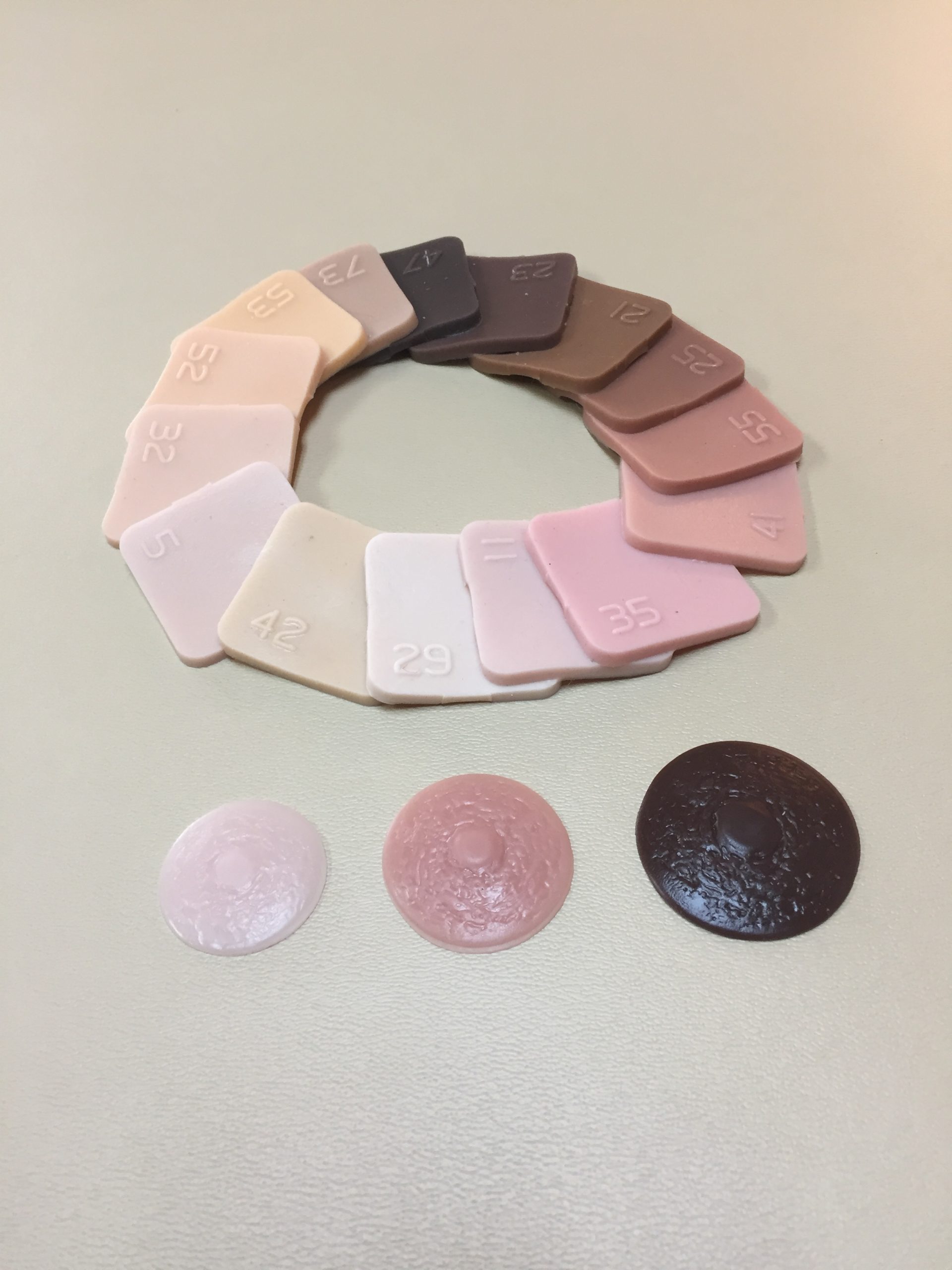 An assortment of about a dozen color sample squares are laid out in an overlapping circle to display the various skin colors available. Three nipple prosthetics of various colors and sizes are also laid in front.